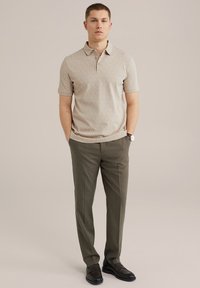 Camisa polo de color beige con un patrón, con cuello y mangas cortas; combinada con pantalones de vestir verde oliva y zapatos de vestir negros, en posición de pie.