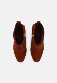 Bottes cheville en daim bordeaux avec un bout pointu, une texture lisse, une languette de tirage et un détail de fermeture éclair dorée sur le côté.