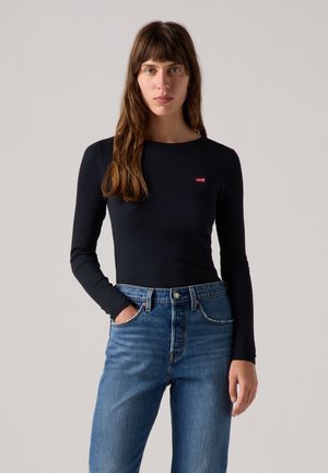 Femme aux longs cheveux bruns portant un t-shirt noir à manches longues avec un petit logo rouge et un jean bleu taille haute, debout sur un fond uni.