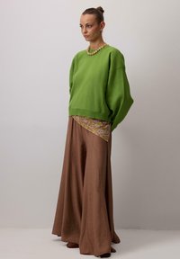 Zelen pulover velikih dimenzij z dolgimi rokavi, nošen preko vzorčastega rjavo-rumenega topa, kombiniran z rjavimi širokimi hlačami in rjavimi čevlji.