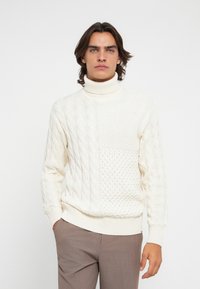 Mann mit schulterlangem Haar, der einen weißen strukturierten Rollkragenpullover und braune Hosen trägt, steht vor einem schlichten weißen Hintergrund.