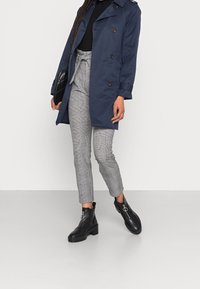 Trench-coat bleu marine en tissu brillant, associé à un pantalon à carreaux gris et des bottines noires avec un accent circulaire sur la fermeture éclair.
