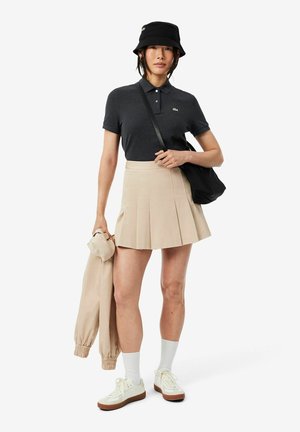 Jonge vrouw met een zwarte bucket hat, donker poloshirt, beige geplooide rok, witte sokken, sneakers, houdt een beige jas en een zwarte schoudertas vast.