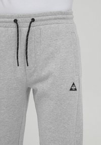 Gray sweatpants made from soft fabric, featuring a black drawstring, side pockets, and a black triangular logo patch on the left leg.