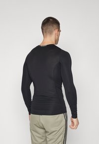 Mand iført en tætsiddende sort langærmet Techfit skjorte og beige bukser med sorte striber, med ryggen mod kameraet foran en ensfarvet baggrund.