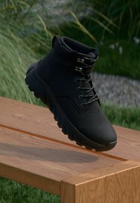 Scarpe da trekking nere con suola in gomma robusta e纹ata, passanti per lacci e design alto alla caviglia, ambientate in uno sfondo naturale.