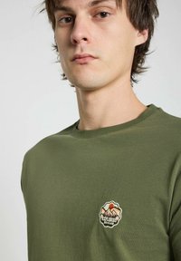 T-shirt en coton vert olive avec un col rond et un petit logo brodé représentant une montagne sur le côté gauche de la poitrine.