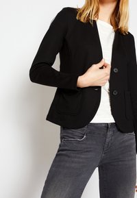 Blazer negro a medida con cierre de dos botones, que presenta un diseño de canalé texturizado y bolsillos en la parte frontal, combinado con jeans grises.