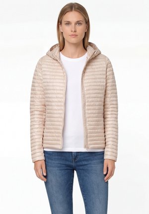 Vrouw draagt een beige gewatteerde jas met capuchon over een wit shirt en een blauwe spijkerbroek, staand tegen een witte achtergrond.