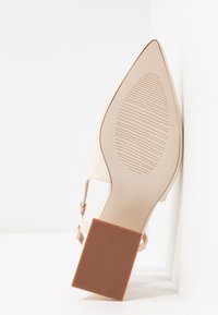 Chaussure beige à bout pointu avec semelle texturée et talon carré marron, positionnée à la verticale contre une surface blanche.