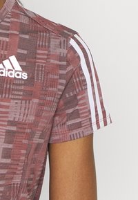 Maglietta sportiva rossa con motivo, maniche corte, con dettagli a tre strisce bianche sulle spalle e logo Adidas sulla parte frontale.