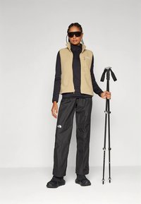 Beige fleecväst över en svart långärmad tröja, kombinerad med svarta vattentäta byxor och svarta vandringsskor, håller i vandringsstavar.