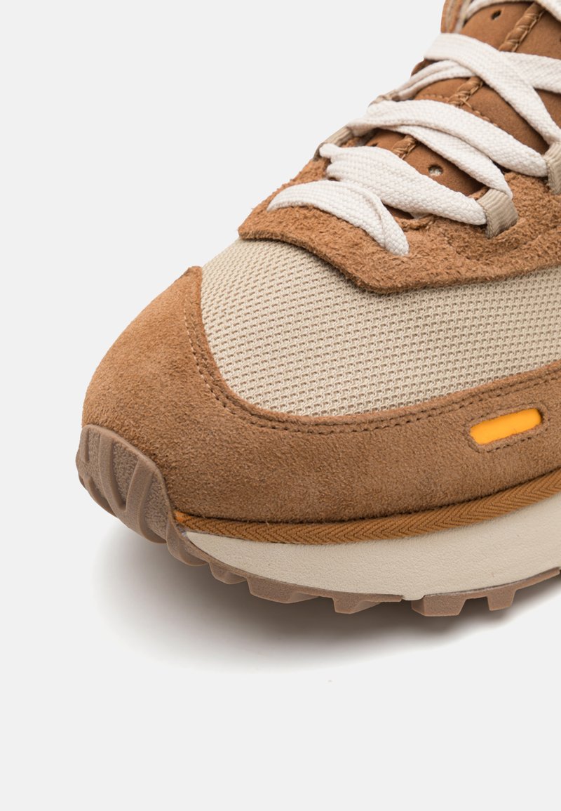 Brązowe zamszowe sneakersy z beżową siateczką, białymi sznurowadłami, teksturowaną powierzchnią, pomarańczowym akcentem i gumową podeszwą z wzorzystym bieżnikiem.