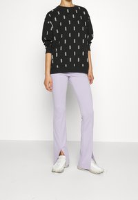 Felpa nera con motivo di testo ripetuto bianco, abbinata a pantaloni a palazzo lilla con spacchi laterali, e sneakers bianche con accenti beige.