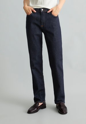 Person trägt gerade dunkelblaue Jeans und dunkelbraune Leder-Loafers, steht mit Händen in den Taschen vor einem einfarbigen hellen Hintergrund.