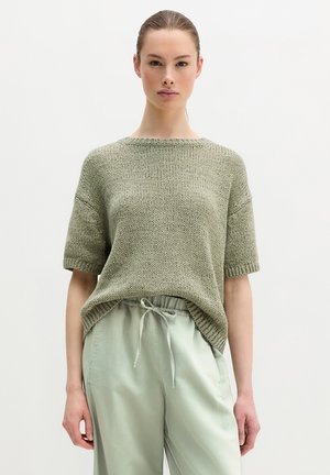 Kobieta z zaczesanymi do tyłu włosami, ubrana w luźny, krótki sweter z dzianiny i jasne spodnie z troczkami, na jednolitym tle.