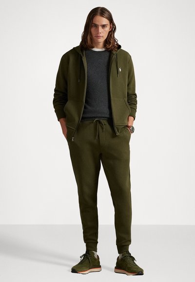 Λαδί φούτερ και αντίστοιχοι joggers φτιαγμένοι από μαλακή σύνθεση βαμβακιού. Διαθέτουν κορδόνια, τσέπες και στενή εφαρμογή. Μαύρα αθλητικά παπούτσια με λεπτομέρειες.