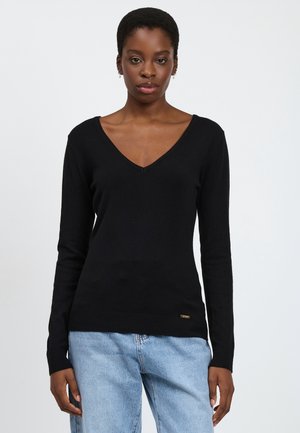Suéter negro de cuello en V con mangas largas, dobladillo acanalado, textura suave y sutil logo de la marca en el lado inferior izquierdo. Combinado con jeans de mezclilla azul claro.
