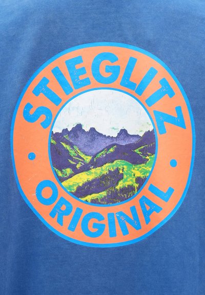 Mėlyna marškinėliai su apvaliu piešiniu, kuriame užrašyta "STIEGLITZ ORIGINAL", vaizduojančiu kalnus ir žalius kalnus ryškiomis spalvomis.