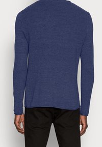 Pull en tricot bleu à manches longues avec une finition texturée, encolure arrondie et ourlet prolongé, associé à un pantalon noir.