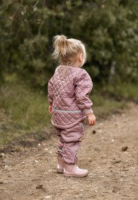 Lyserød quiltet jakke og matchende bukser med elastiske manchetter; kombineret med lyserøde gummistøvler. Scenen viser et barn, der går på en jordsti.