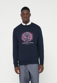 Bărbat îmbrăcat cu un hanorac bleumarin cu logo-ul roz Timberland, o cămașă albă dedesubt și pantaloni albaștri, stând în fața unui fundal alb simplu.