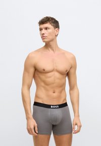 Γκρι μποξέρ με μαύρη ζώνη μέσης που φέρει το λογότυπο "BOSS". Λείο ύφασμα, εφαρμοστή γραμμή και μήκος μέχρι το μέσο του μηρού.