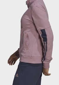 Roze jas met rits en een gestructureerde afwerking, voorzien van marineblauwe drie-streep accenten op de mouwen en een zijzak voor extra functionaliteit.