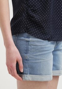 Main gauche reposant sur le côté, portant un short en jean bleu clair retroussé et une chemise bleu foncé à petits pois blancs.