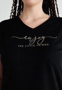 Μαύρο T-shirt με V λαιμόκοψη φτιαγμένο από υφή υφάσματος, που διαθέτει χρυσό καλλιγραφικό κείμενο "απόλαυσε ΤΑ ΜΙΚΡΑ ΠΡΑΓΜΑΤΑ" μπροστά.