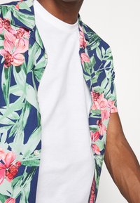 Kurzärmeliges Hemd mit Knöpfen in Blau mit floralem Muster, grünen Blättern und pinkfarbenen Blumen. Darunter ein weißes T-Shirt mit Rundhalsausschnitt.