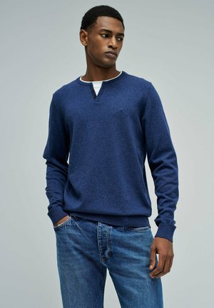 Joven con cabello corto, vestido con un suéter henley azul sobre una camiseta blanca y jeans azules, de pie con una mano en el bolsillo.