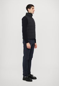 Black quilted vest with high collar, worn over a navy long-sleeve shirt, paired with navy pants and black lace-up shoes.