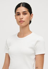 Balta juostuota t-shirt marškiniai su trumpomis rankovėmis ir apvaliu iškirpimu. Lygus paviršius, prigludusi forma, su subtilia siūlės detale ant pečių.