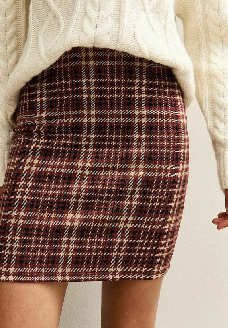 Mini-jupe moulante à motif à carreaux rouge, noir et beige portée sous un pull en maille torsadée crème.