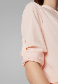 Blouse rayée de couleur pêche clair avec des manches à rouleaux à boutons et une texture douce, dotée d'un col et d'un léger éclat.