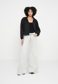 Melna bomber stila jaka, pieguļoša, ar rāvējslēdzēju; kombinēta ar baltām izplatītām džinsa biksēm, gaisīgas krāsas, mīksta tekstūra. Modelis pozē pret vienkāršu fonu.