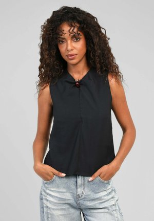 Femme aux cheveux bouclés portant un haut noir sans manches avec un bouton décoratif et un jean bleu clair, debout les mains dans les poches.