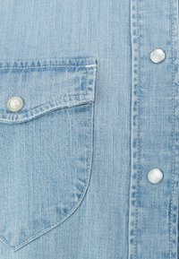 Svijetloplava traper košulja s džepom na prsima, perlasta dugmad i suptilna vertikalna tekstura.