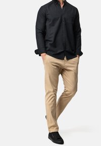 Chemise noire boutonnée avec une poche, associée à un pantalon beige et des baskets noires. La tenue a un style décontracté mais soigné.