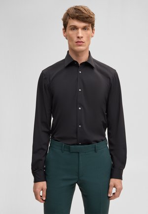 Junger Mann steht, trägt ein schwarzes Hemd mit Knöpfen und dunkelgrüne, maßgeschneiderte Hose vor einem schlichten hellen Hintergrund.