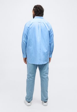Homme aux cheveux tressés portant une chemise bleu clair à manches longues, un jean bleu clair et des baskets blanches, debout de dos.