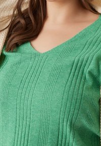 Grüner Strickpullover mit V-Ausschnitt, der über eine vertikale Rippstruktur und weichen Stoff verfügt. Das Design beinhaltet erhabene Linien für zusätzliche Details.