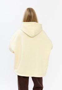 Gele oversized hoodie met een grote capuchon, geribbelde zoom en een zachte textuur. Model draagt een paarse corduroy broek en staat met de rug naar de camera.