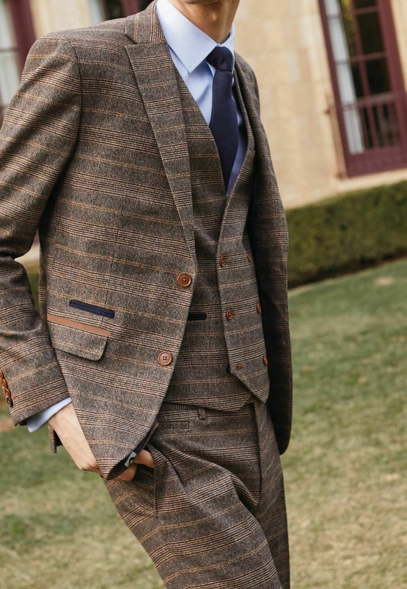 Bruin geruit driedelig pak gemaakt van gestructureerde stof met bruine knopen en blauwe accenten. Witte overhemd en navy das maken de look compleet.