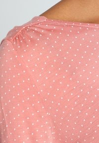 Ljusrosa kortärmad topp gjord av mjukt tyg, med ett strukturerat mönster av små vita trianglar som är jämnt fördelade över ytan.