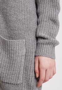 Szary dziany sweter z teksturowanym wzorem, z boczną kieszenią i prążkowanymi mankietami. Tkanina wydaje się miękka i gruba.