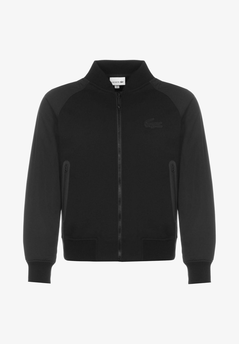 Veste noire à fermeture éclair avec col et poignets côtelés, deux poches latérales zippées et un logo discret brodé sur la poitrine.