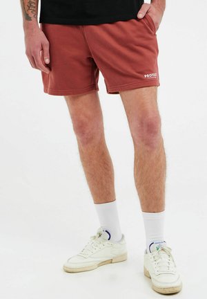 Rote Sweathorts aus weichem Material, ausgestattet mit Seitentaschen und einem Logo. Kombiniert mit weißen Socken und weißen Sneakers mit blauen Akzenten.