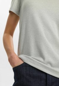 Tricou drept, de culoare gri deschis, cu mâneci scurte, având o croială lejeră și băgat în blugi negri, evidențiind textura moale și luciul subtil.